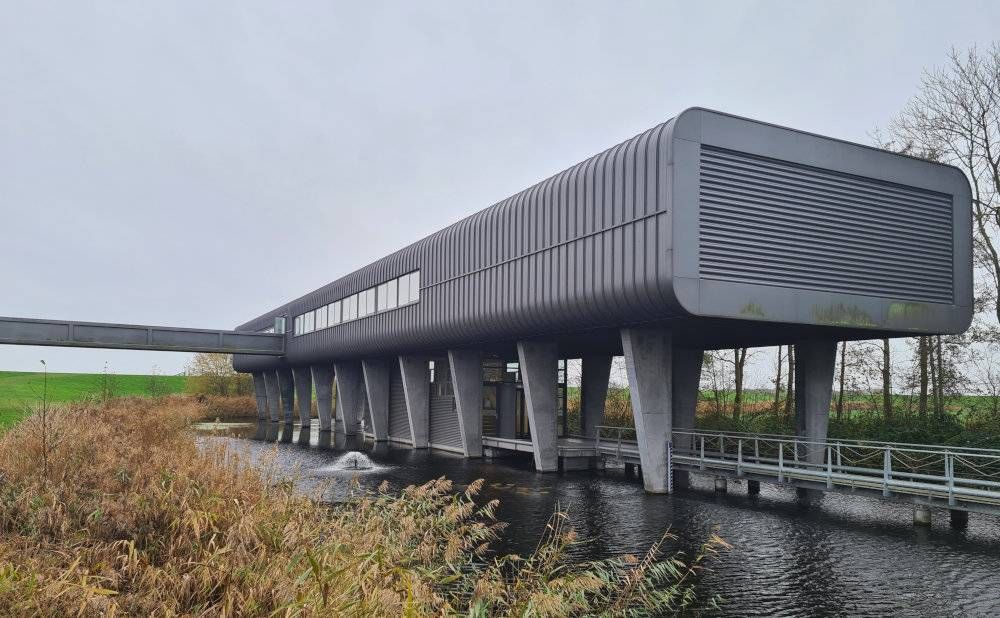 A very modern building - gray and long and narrow, standing above water on two rows of concrete legs.