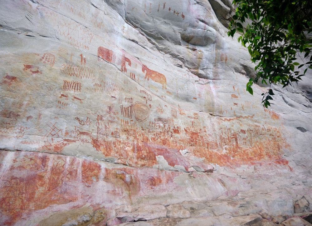 A rock face covered in small pictures of people and animals in reddish colors.