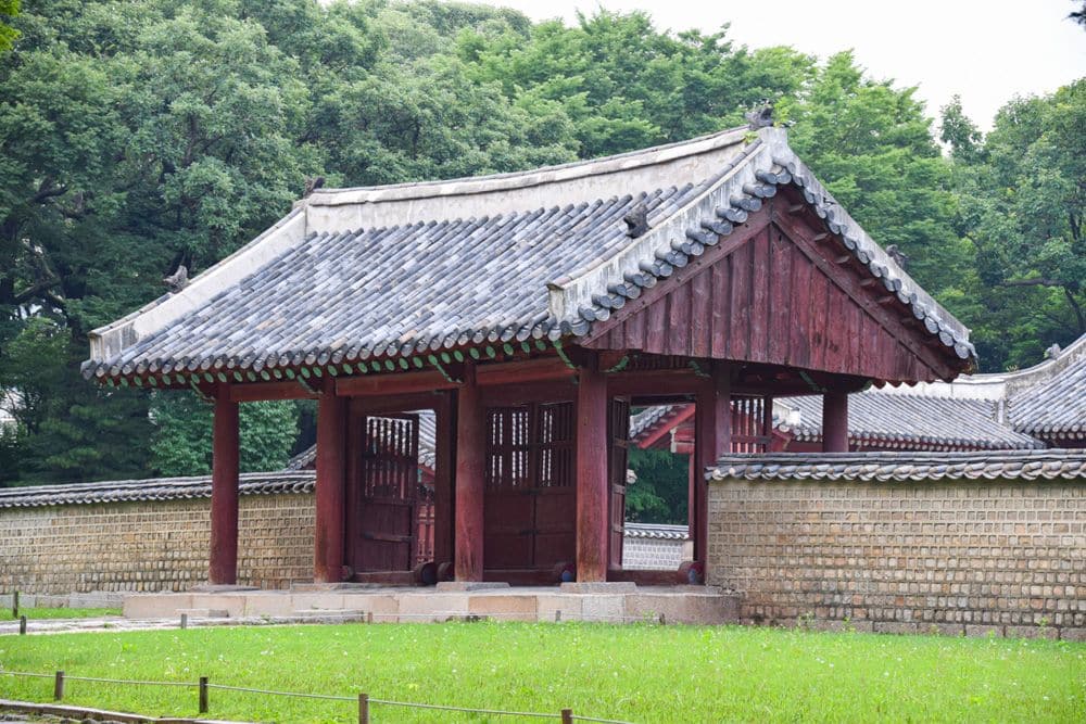 A small and simple gate in a wall: peaked tile roof over an open story supported by pillars, painted dark red.