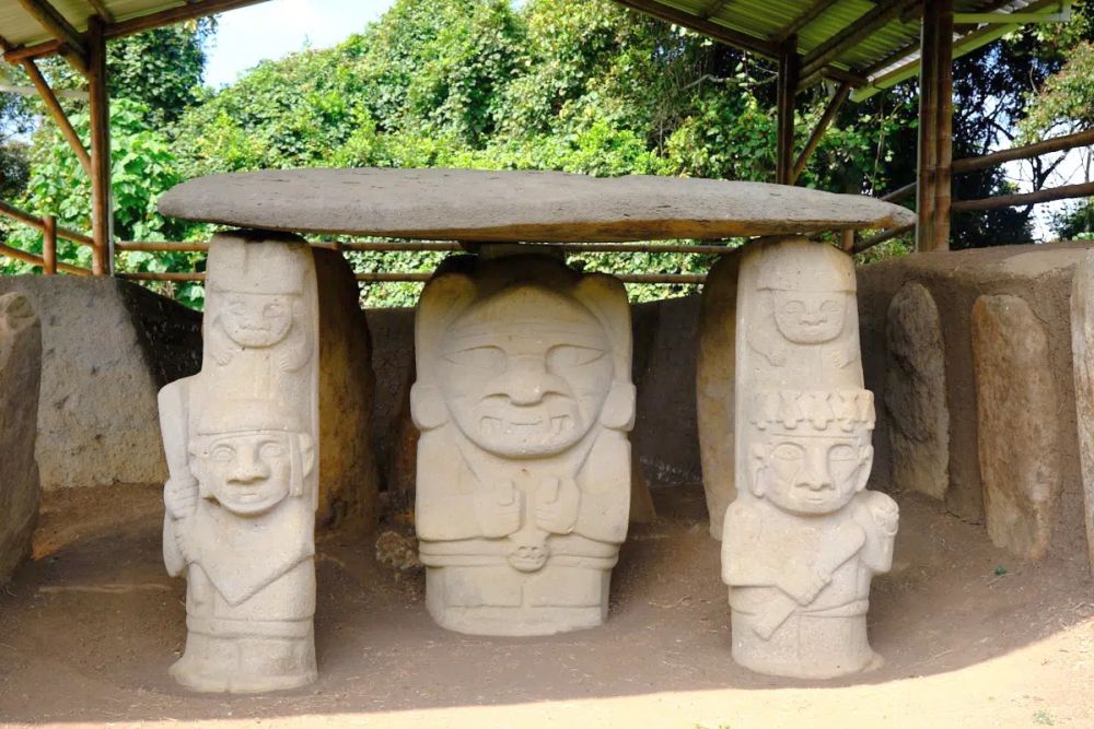 Three statues, with more behind them that are barely visible, support a large flat stone, forming a table. The statues have human faces wearing headresses and carrying clubs of some sort.