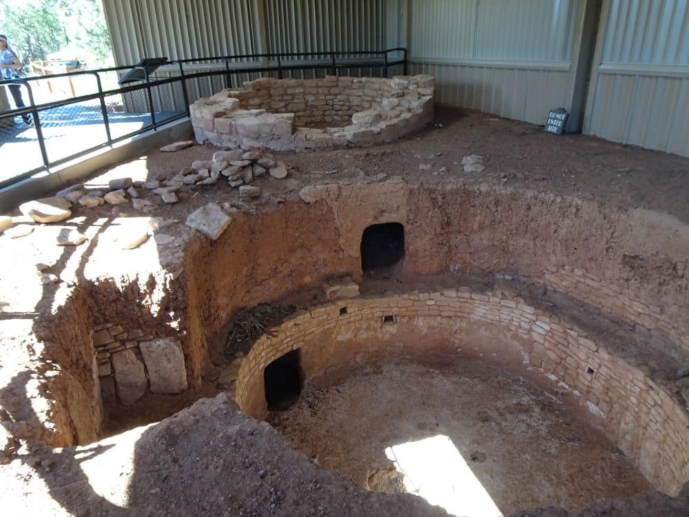 A dug-out area of ground seems to be the excavation of a kiva, with round walls. A modern roof is above it and it is fenced off from people.