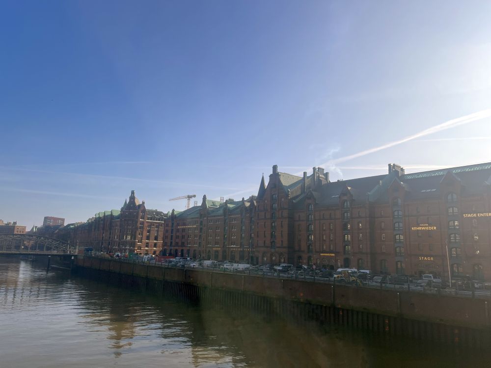 View across a canal or river to a row of very large brick buildings along the bank.