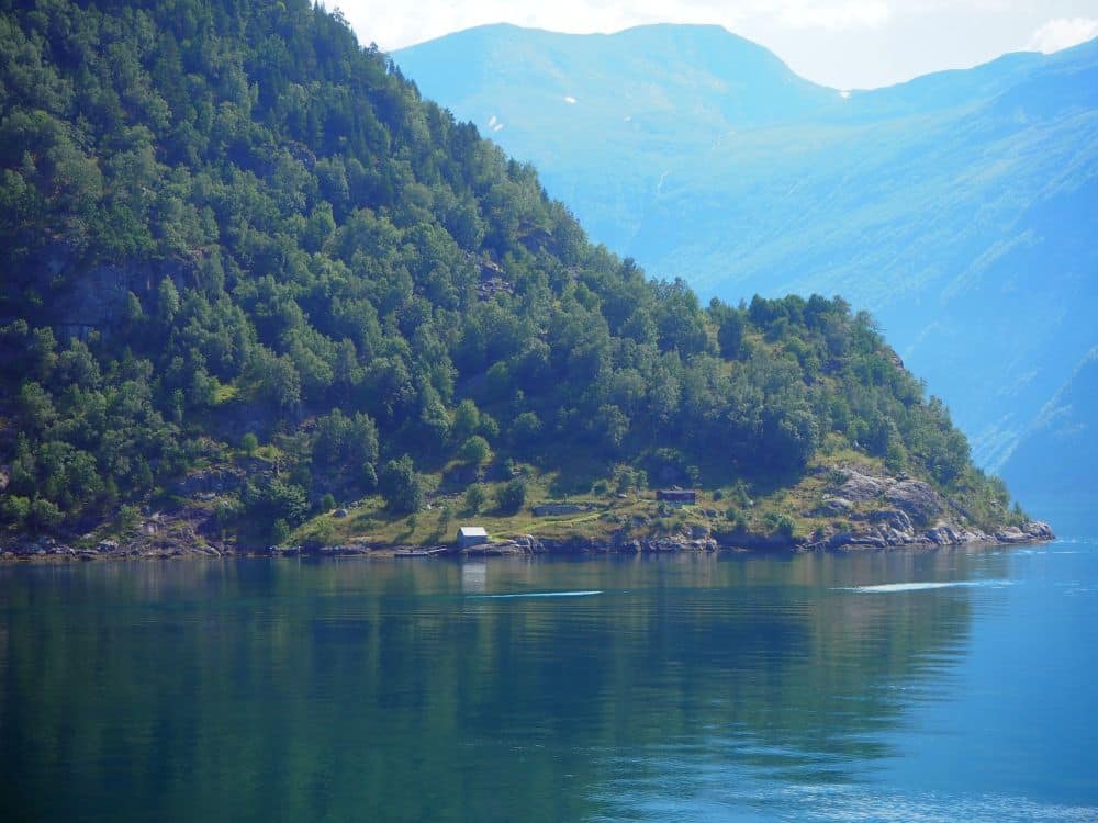 Between a steep mountainside and the edge of the water, a small cottage in a small clearing, with a boathouse on the edge of the water. A large mountain looms behind.