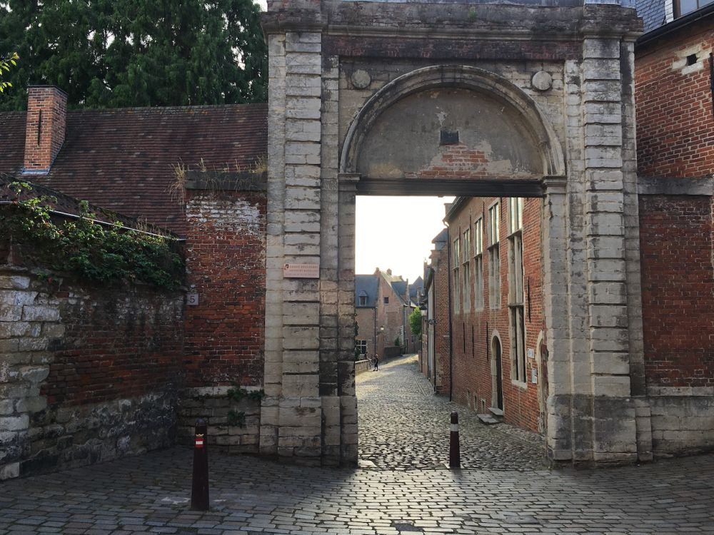 An arched entryway with open door allows a view down a cobblestone street lined with brick houses.