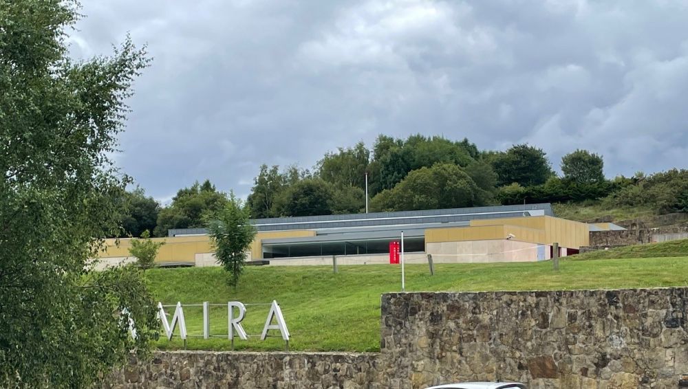 A long low modern building with a grassy lawn. A sign on the near end of the lawn on top of a stone wall is partly obscured by a tree, so only the letters "MIRA" are visible.