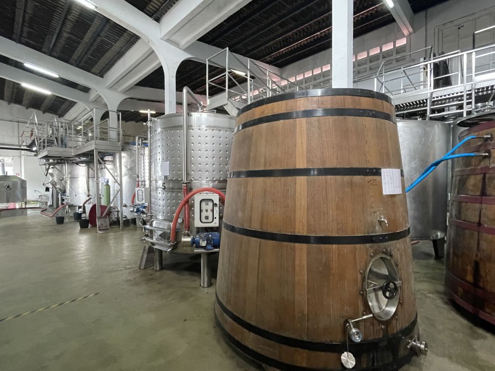 Inside a winery, a row of large shiny metal vats, and the nearest vat is wood, like an extra large wooden barrel.