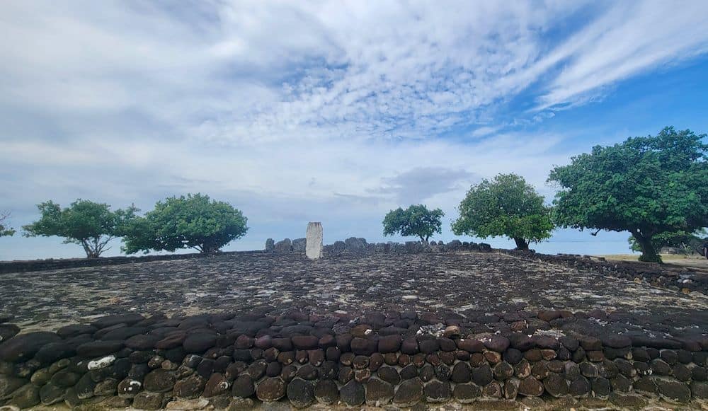 A field paved with dark volcanic rocks, trees along its edge, a stone wall in front, and larger stones at the far end.