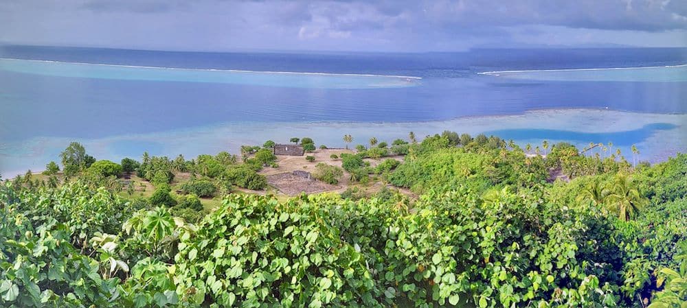 A view across a green area of land with a clearing of a marea on it, along with a low building. Beyond the land, far below, is blue sea, where the reef channel is visible.