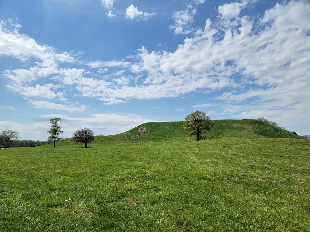 A flat grassy field with, at a distance, a large grass-covered mound, flat on its top.