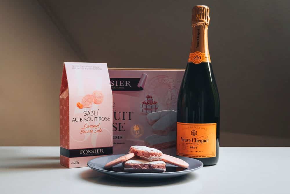 A pale pink box and a pale pink bag of biscuits with, in front of them, a plate of pink biscuits and a bottle of Veuve Clicquot champagne.