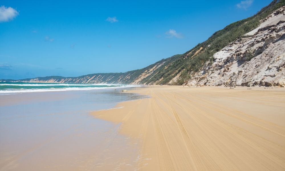 looking down a white-sand beach, hills rising from it with scrubby green plants over white rock.