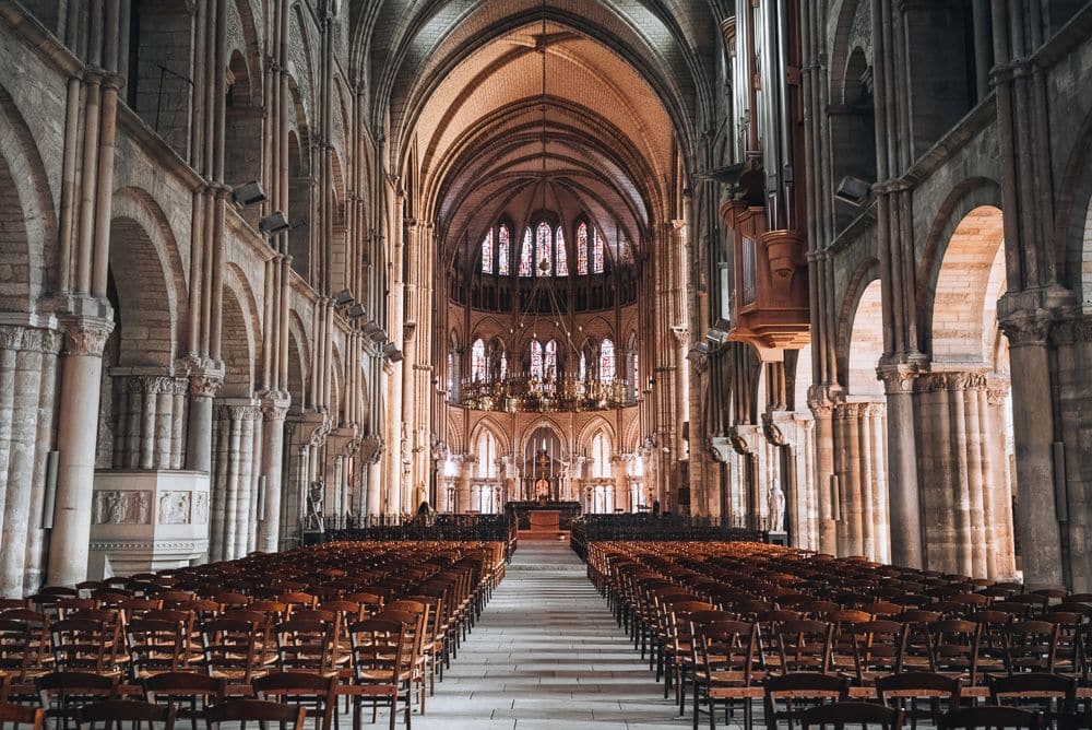 A large view down the center of the church with Romanesque archways along the sides and a gothic arched ceiling. The altar is far in the distance, with stained-glass windows behind it.