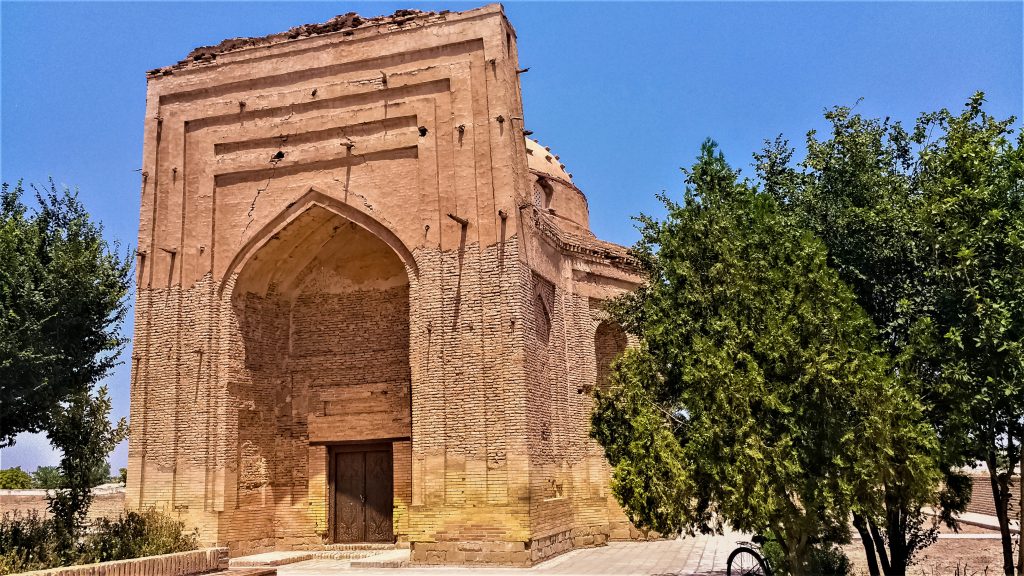 A building brick, partly stuccoed, with a very large entry archway: pointed, but then the wall around it is rectangular, with decorative elements above the archway. Behind the entance, part of a dome can be seen, not much higher than the archway.