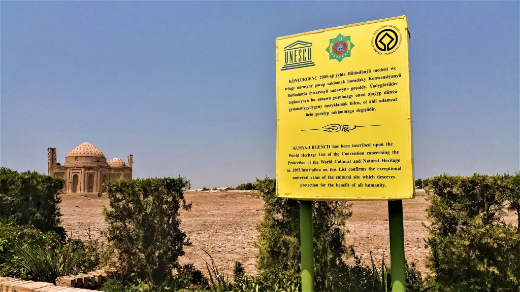 A sign at the entrance to Kunya-Urgench says essentially that it's a UNESCO site and why. Behind the sign, a barren and stony stretch of ground and, in the distance, a mosque or mausoleum.
