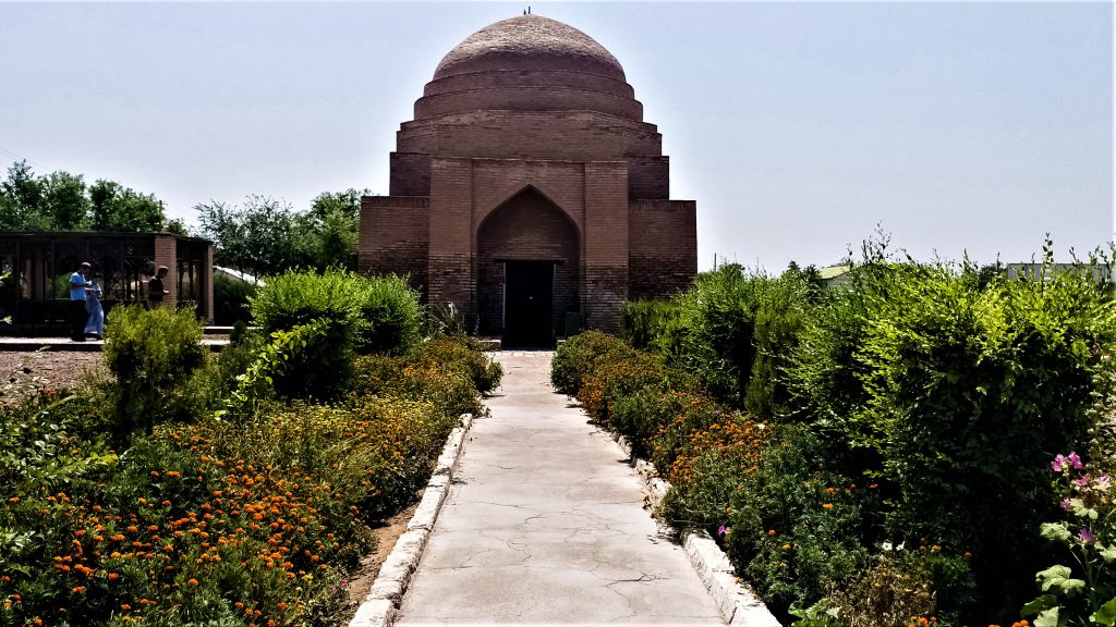 A path straight ahead with bushes on either side, leading to a large domed building with an arched entryway.