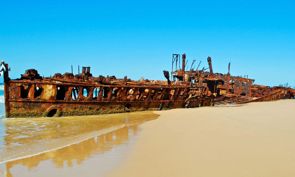 The rusting carcass of a ship sits half on the white sand and half in the shallow water.