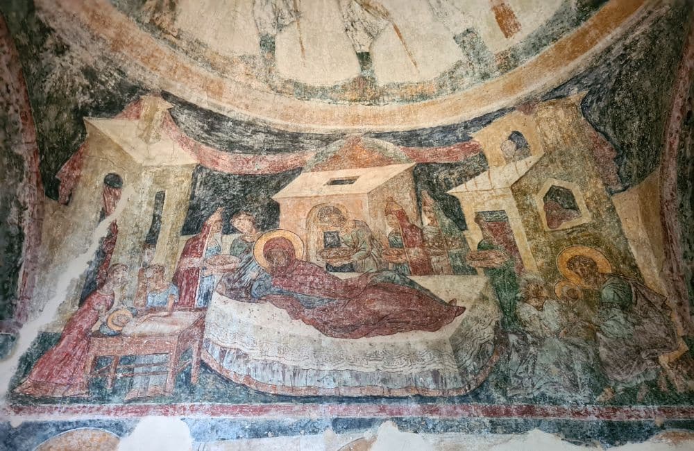The fresco is faded but still colorful. It seems to depict the birth of Jesus - a woman with a halo lies in the center, and a baby with a halo is nearby on a small table, with 2 women tending to it. The 3 magi and others hold gifts around her bed. 