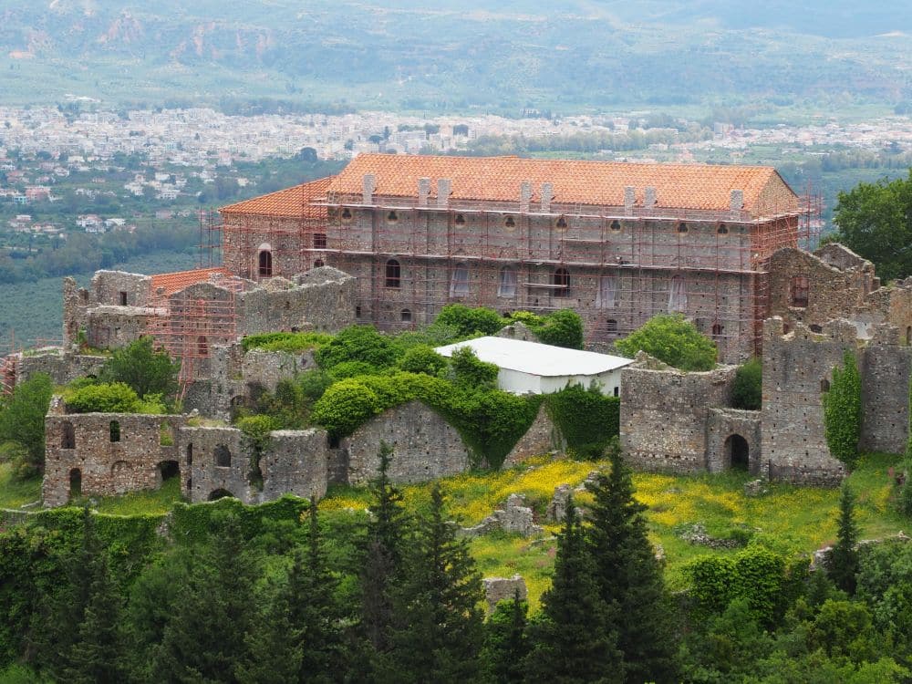 A building of just a few stories, quite long, stone with a red roof. It is surrounded by stone ruins.