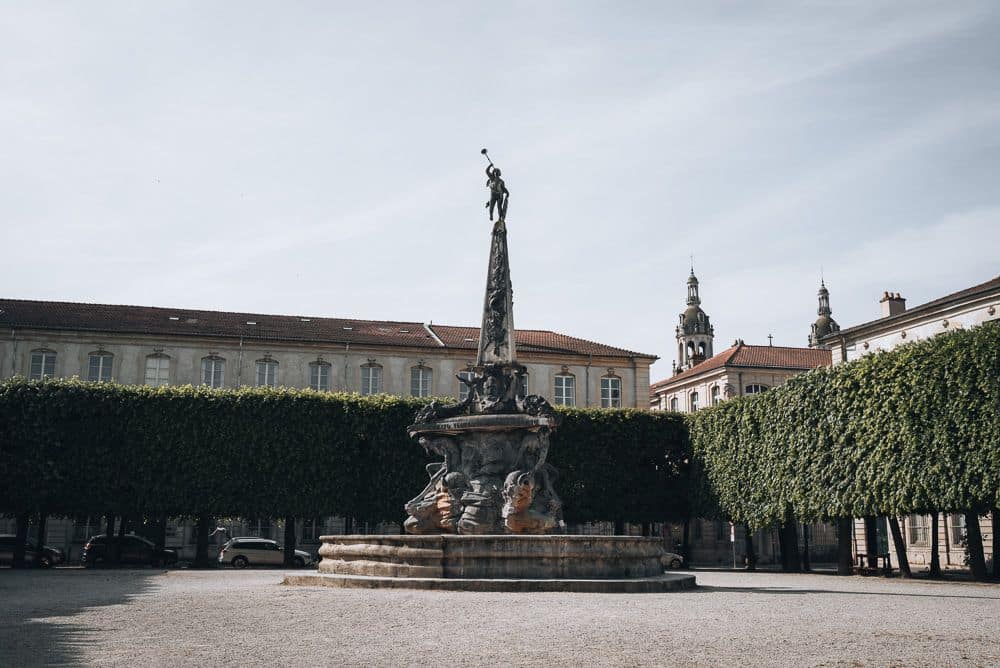 A tall, elegant statue in an open plaza. Very neatly trimmed trees surround the plaza, and the buildings are partly visible above the trees.