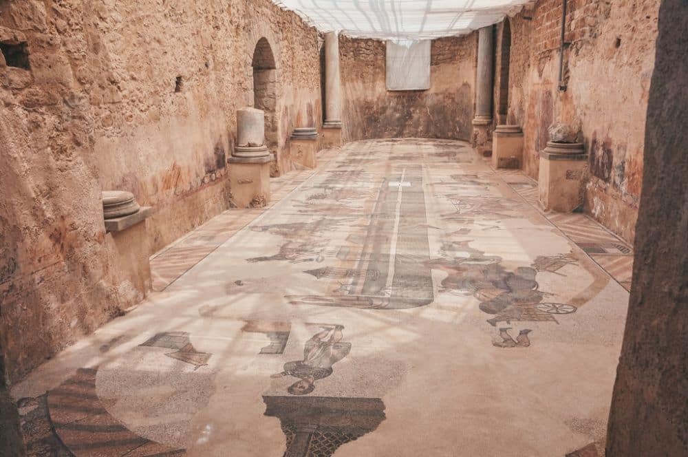 View down a long narrow room with stone walls and a glass roof. The floor is covered in a mosaic with images of people and animals.