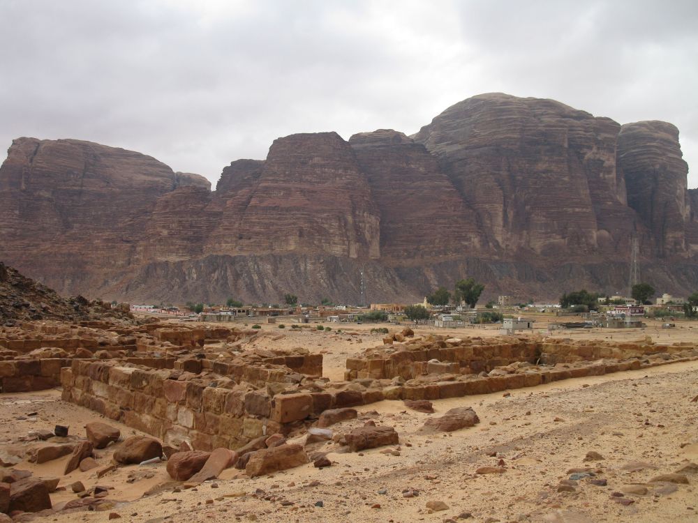 Large cliffs show red sedimentary layers and, in the foreground, the ruins of an ancient building and in the middle distance, some low modern buildings.