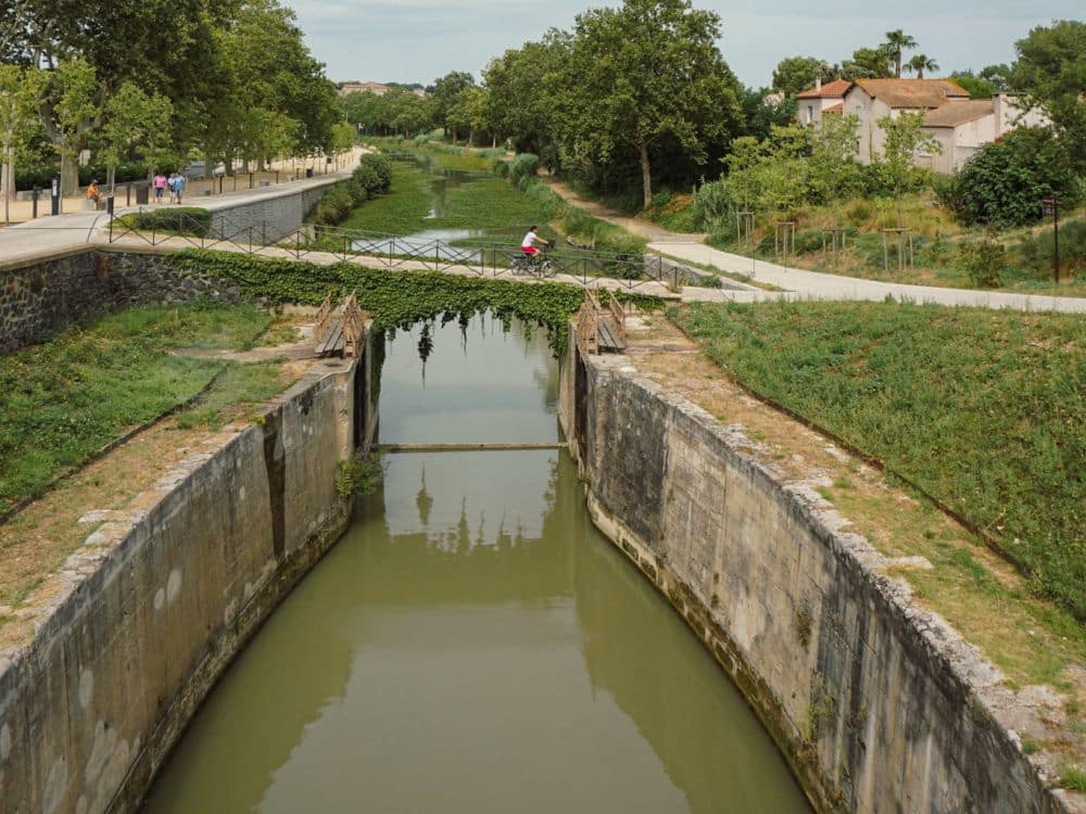 Looking straight down the Canal du Midi, a small bridge crosses it, with a bicycle passing over it.