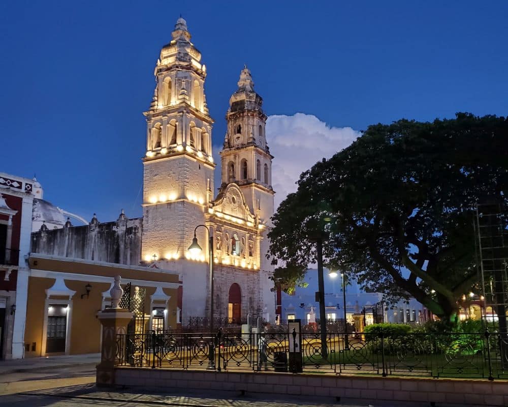 An evening shot on the town square, which is partly visible with some trees on the right. Facing the square is the cathedral, brightly lit up with two tall towers.