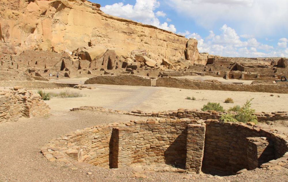 Foreground: a kiva, a round sunken pit. Background: half-ruined buildings.