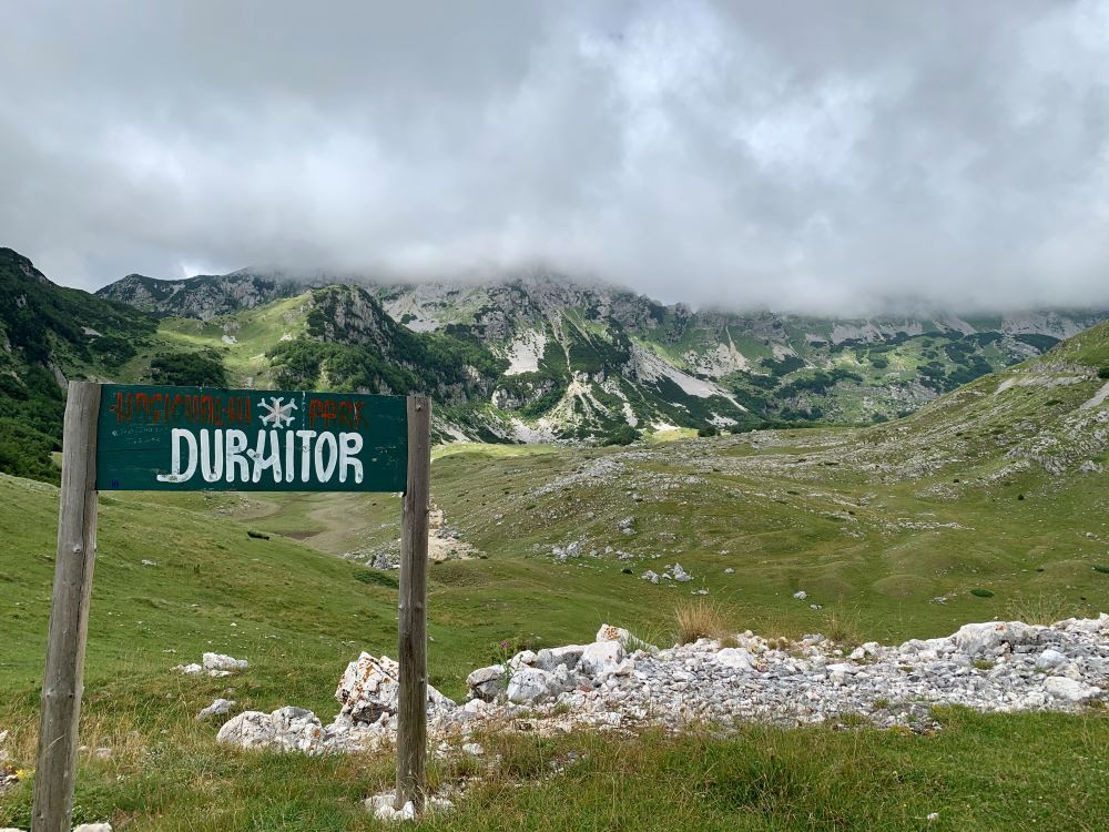 A sign that reads "Durmitor" in front of a view of a mountain ridge topped with clouds.
