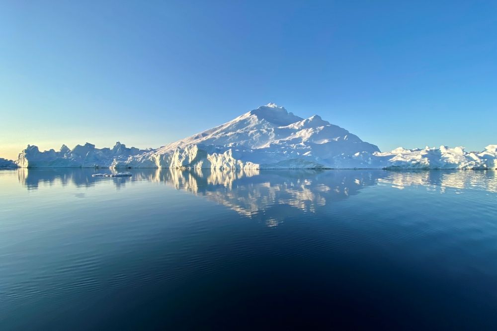 Seen across glassy blue water, a mountain of ice, reflected in the water.