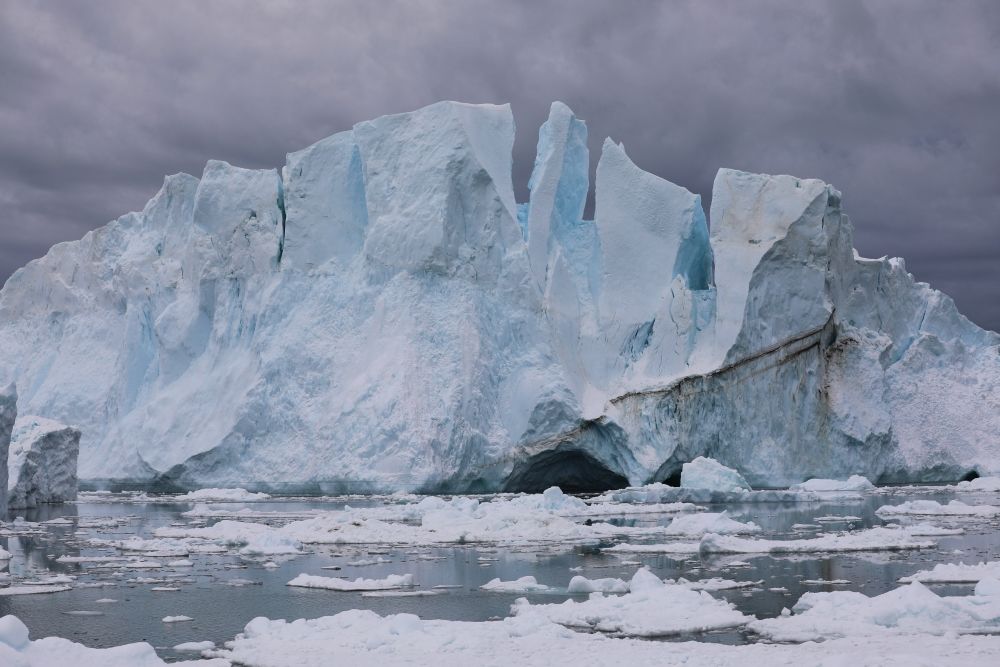 An iceberg, all jagged edges, against a grey sky, and smaller pieces in the grey water around it.