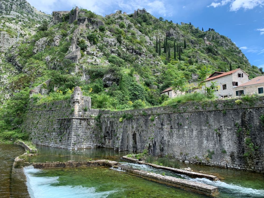 A city wall with water at its base, and a wall snaking its way up a hill behind it.