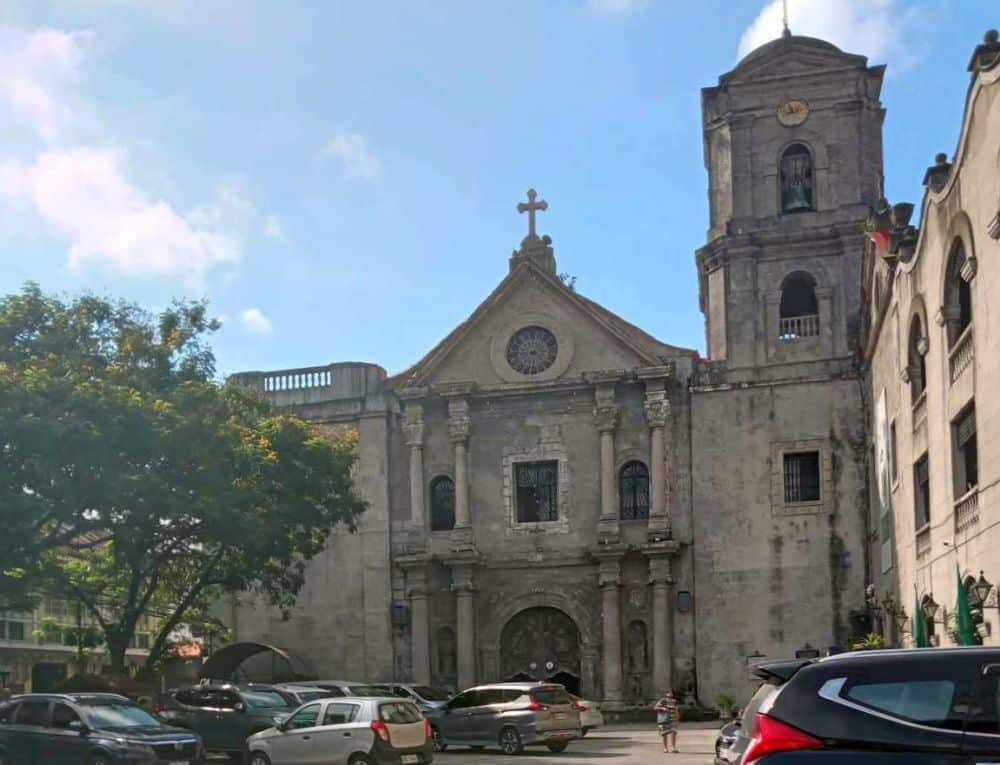 Front facade of the church has classical columns on each of two stories on either side of the central entrance. One tower on the right, almost square but with angled corners.