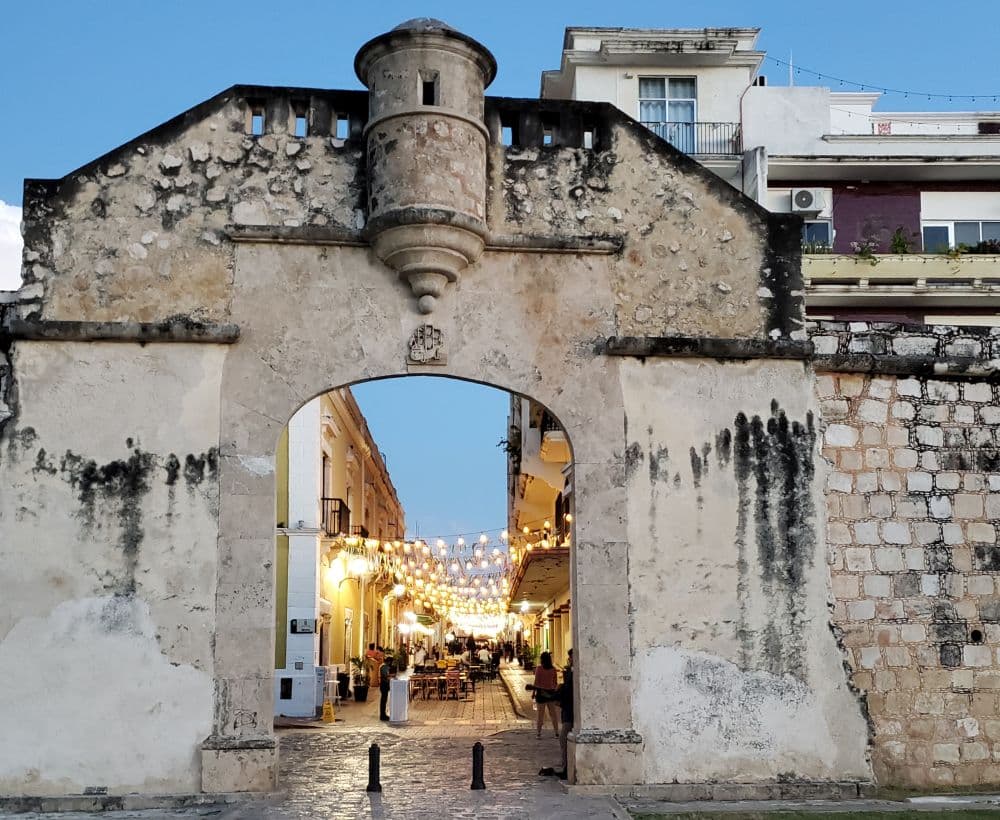 An old colonial-style adobe gateway with a large archway, and beyond it a street lit up with strings of light across the street.