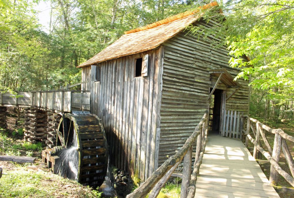 An unpainted wooden building, quite small, with a water wheel extending from its side into a stream, and water falling from the wheel.