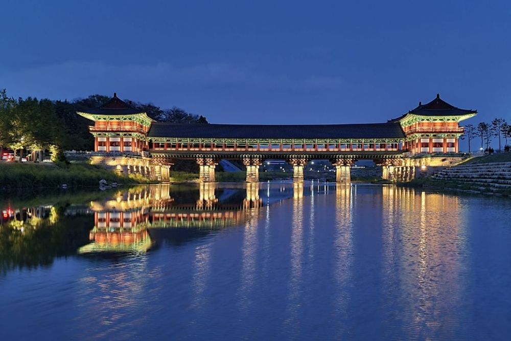 A bridge over a pond or river, with a square tower at each end, all of it lit up in bright colors at night.