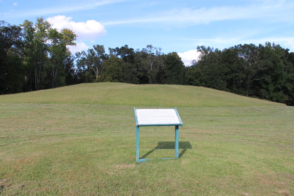 A grass-covered mound at Poverty Point with an informational sign in front of it.