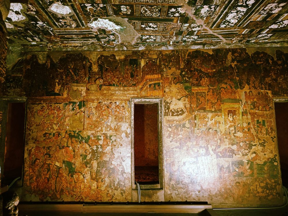 A room with a flat wall with a single door in it. The wall and the ceiling are covered in colorful paintings, though the ceiling is quite damaged.