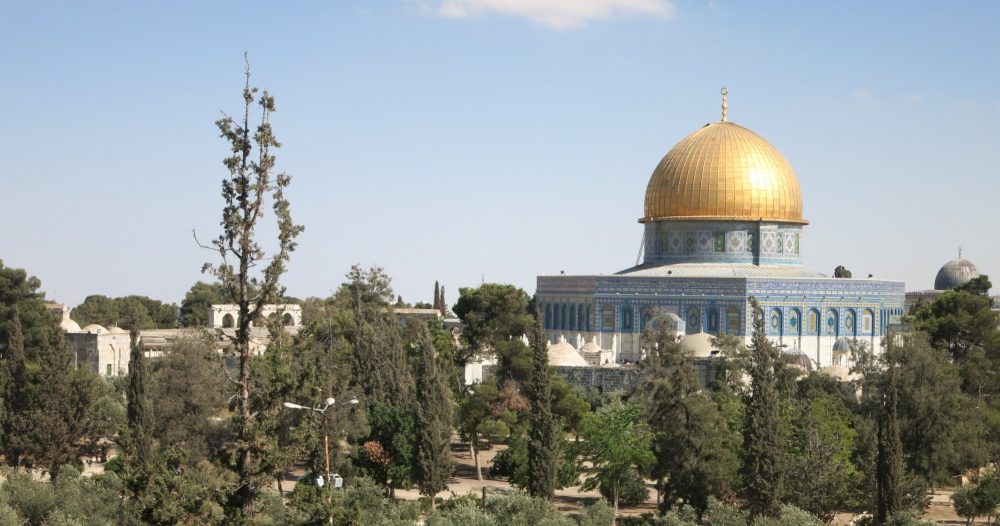 trees in the foreground and behind them a blue-tiled building with a golden dome.