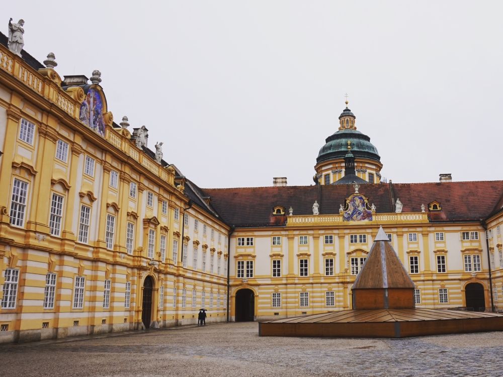 Two sides of Melk Abbey around a courtyard with a fountain or monument in the middle.