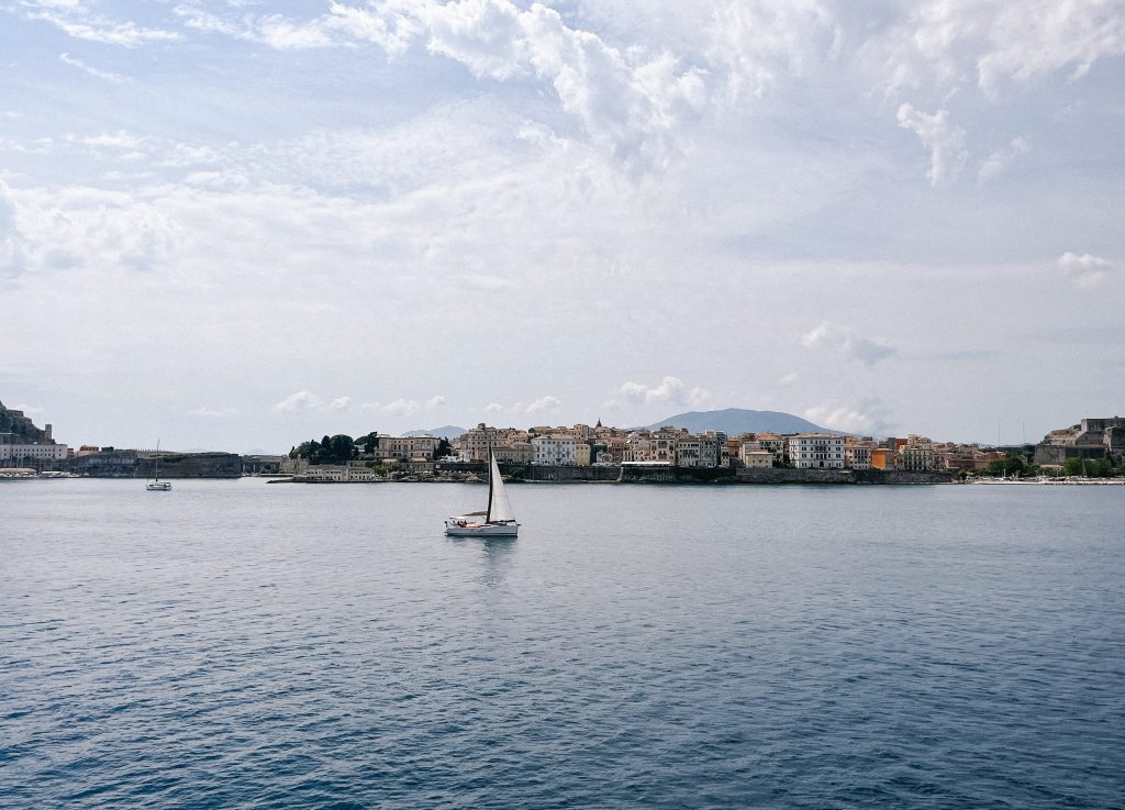 View of Corfu Old Town from the water, with a sailboat in front of the coastline.