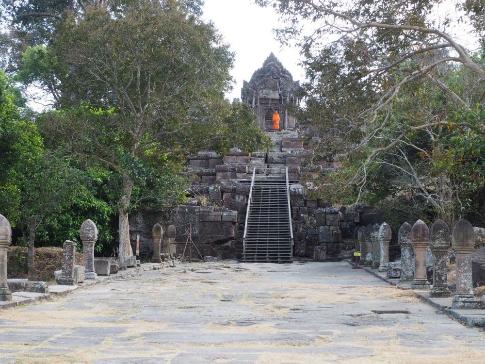 A large flat walkway paved with large stones, short standing stone on both sides, and a small temple up a stairway straight ahead, with a monk in orange at the top.