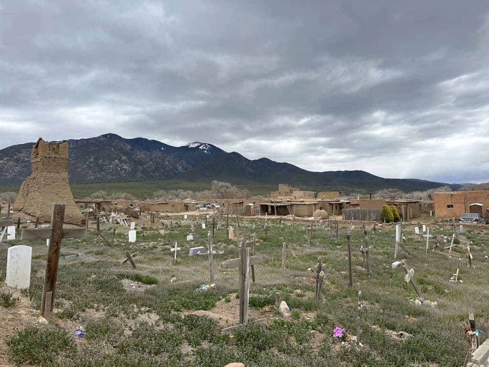 A cemetery with crosses marking the grave and a small stone ruin. Behind that, some adobe buildings.