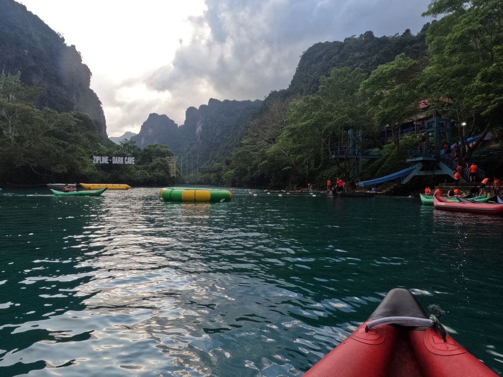 On a river, the point of a kayak in the foreground; mountains on either side of the water. A sign on the hill to the left reads "zipline dark cave" and people baord canoes on the right.