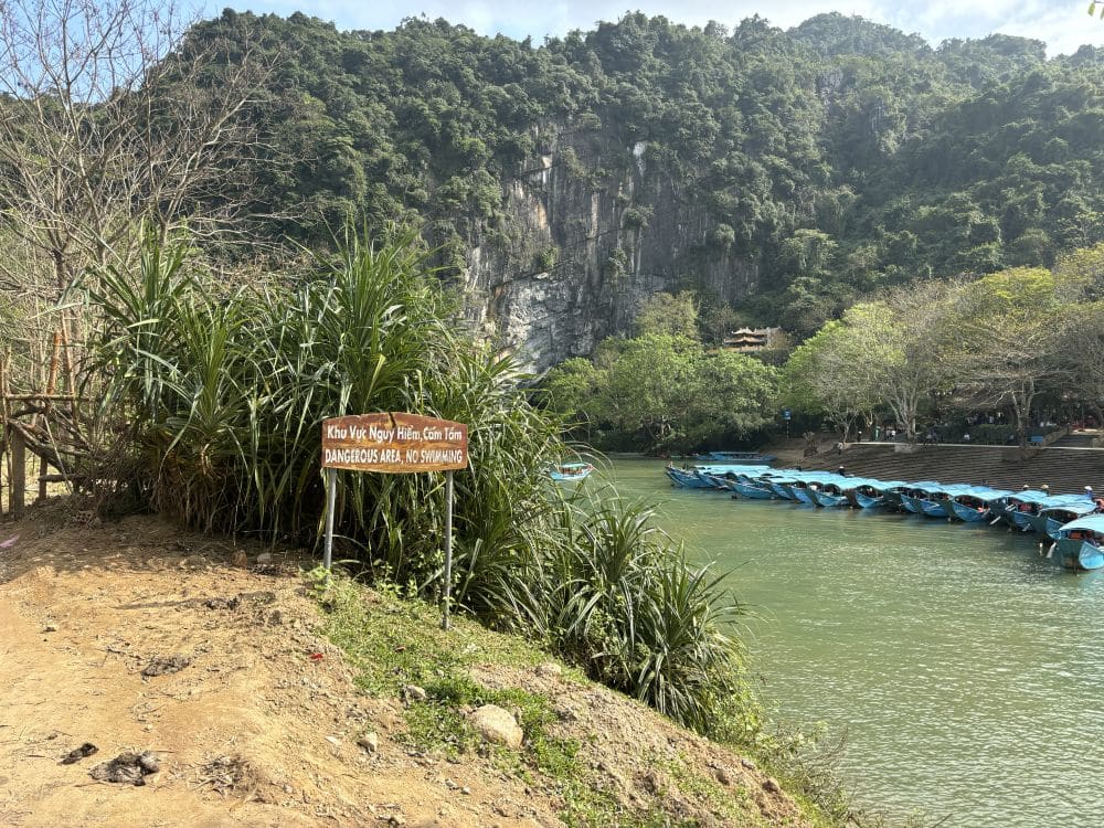 On a riverbank, karst mountains across the way and a row of boats waiting for customers on the river. A sign reads "Dangerous area no swimming."