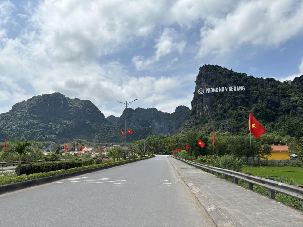 On the road that enters Phong Nha-Ke Bang National Park: karst mountains in the distance, one with a sign with the name of the park. Vietnamese flags along the road.