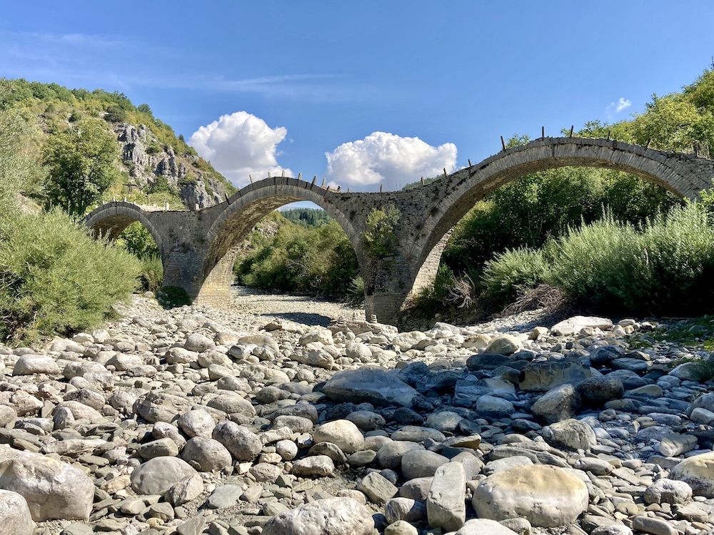 A dry, stony riverbed, with a arched stone bridge spanning it.