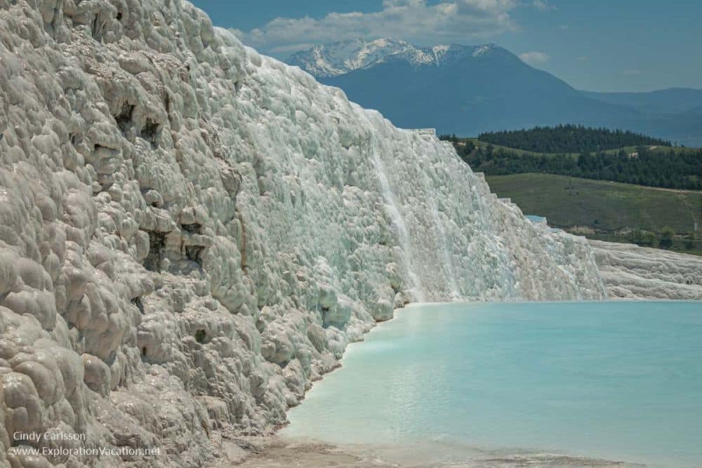 A wall of white minerals next to a very blue pool, with mountains in the distance.