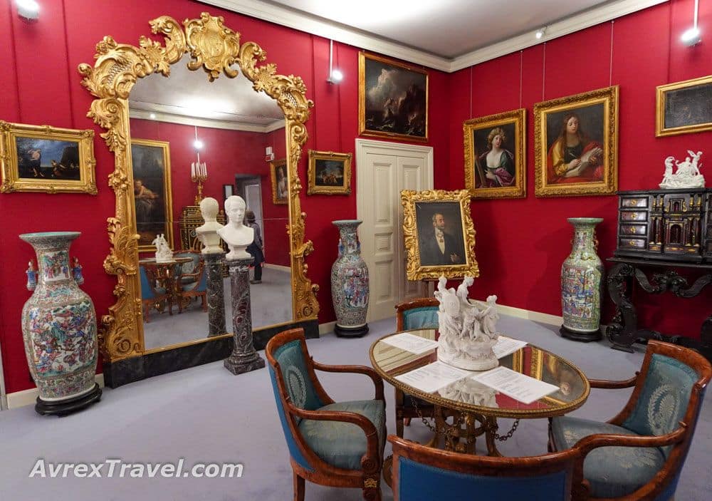 A red-painted room with a very large mirror in an ornate gold frame and portraits on the walls.