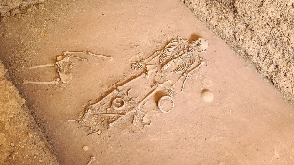 A skeleton, partly protruding from a flat area of reddish soil.